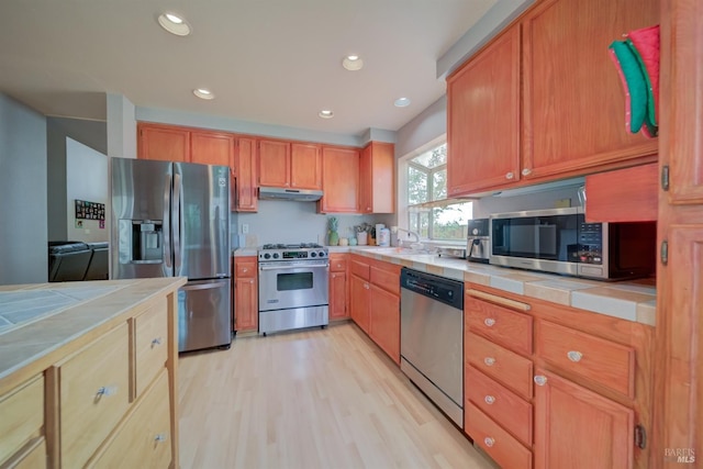 kitchen with tile countertops, under cabinet range hood, appliances with stainless steel finishes, and light wood-type flooring