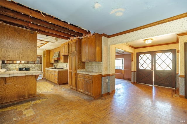 kitchen with ornamental molding, brown cabinets, premium range hood, wallpapered walls, and tile counters