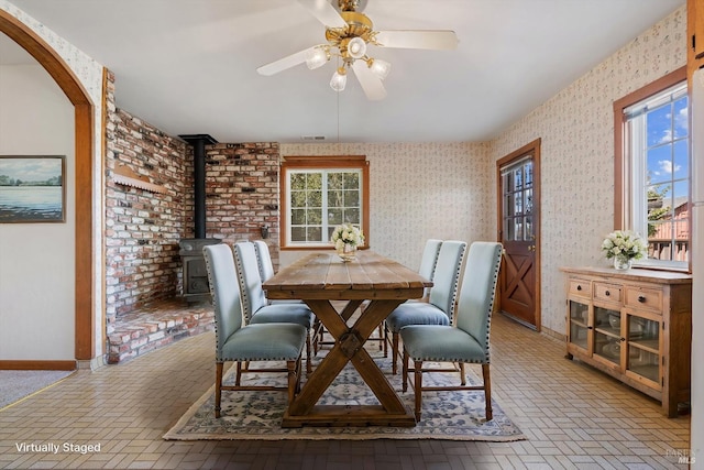 dining space with arched walkways, wallpapered walls, a ceiling fan, brick floor, and a wood stove