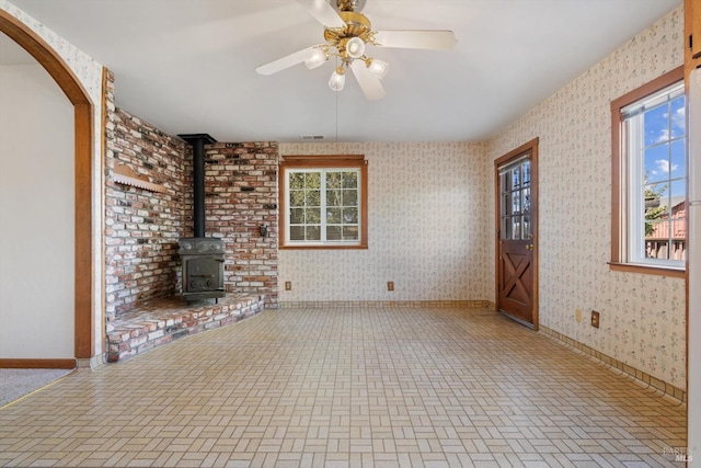 unfurnished living room with baseboards, wallpapered walls, a ceiling fan, brick patterned floor, and a wood stove