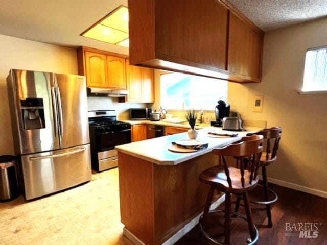 kitchen with light countertops, appliances with stainless steel finishes, a peninsula, under cabinet range hood, and brown cabinets