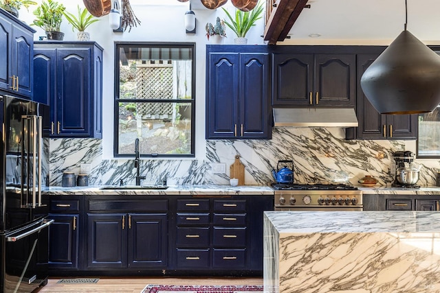 kitchen with blue cabinets, light stone counters, freestanding refrigerator, under cabinet range hood, and a sink