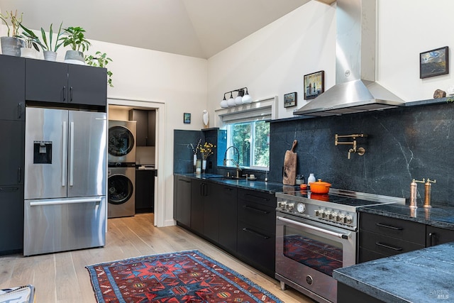 kitchen with a sink, stainless steel appliances, wall chimney exhaust hood, light wood-style flooring, and stacked washer / dryer