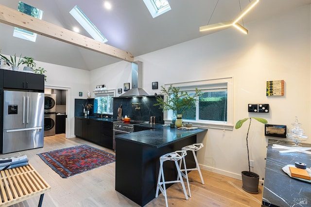 kitchen featuring stacked washer and dryer, stainless steel appliances, wall chimney exhaust hood, high vaulted ceiling, and a skylight