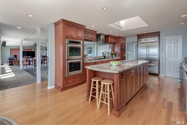 kitchen featuring wall chimney range hood, appliances with stainless steel finishes, a breakfast bar, light wood finished floors, and a skylight