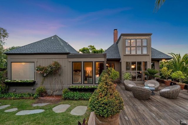 back of property at dusk featuring roof with shingles, a chimney, and a deck