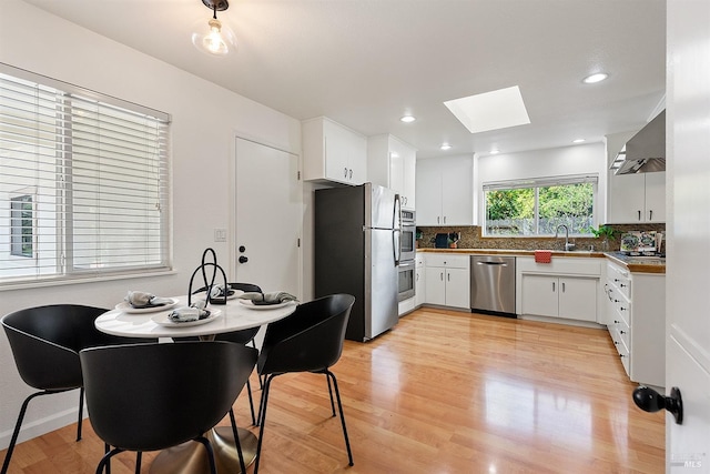 kitchen with wall chimney range hood, backsplash, a skylight, stainless steel appliances, and light wood finished floors