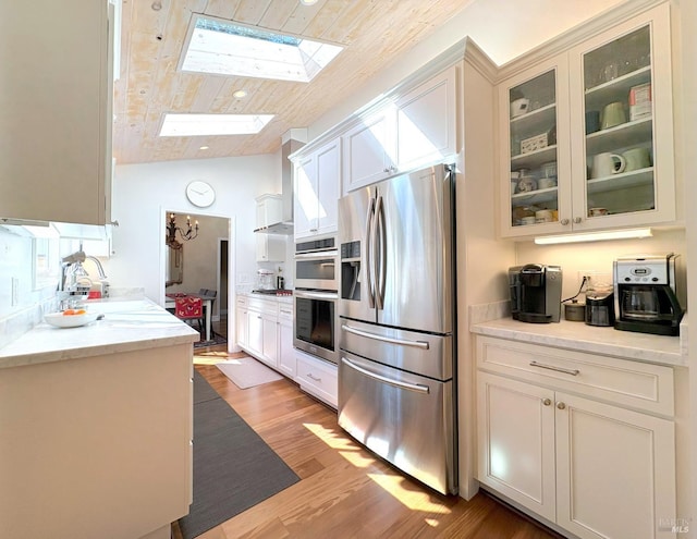 kitchen with wall chimney range hood, lofted ceiling with skylight, glass insert cabinets, stainless steel appliances, and light wood-style flooring