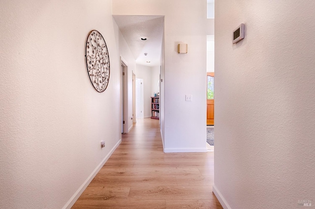 corridor featuring a textured wall, light wood-type flooring, and baseboards