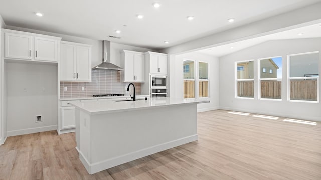 kitchen featuring white cabinets, oven, a sink, wall chimney exhaust hood, and a center island with sink