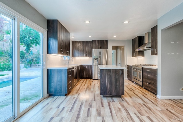 kitchen featuring wall chimney exhaust hood, light wood-type flooring, light countertops, stainless steel appliances, and backsplash