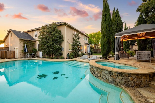 pool at dusk with a pool with connected hot tub, a gazebo, fence, and a patio