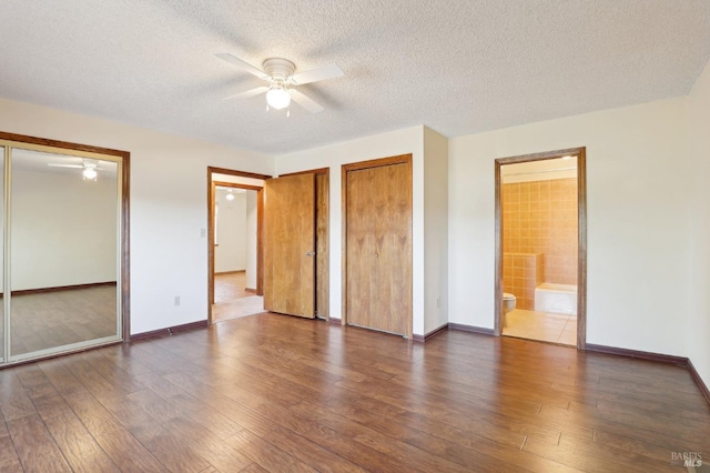 unfurnished bedroom featuring two closets, hardwood / wood-style floors, baseboards, a textured ceiling, and ensuite bath