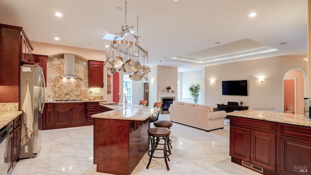 kitchen with arched walkways, wall chimney range hood, appliances with stainless steel finishes, a tray ceiling, and reddish brown cabinets