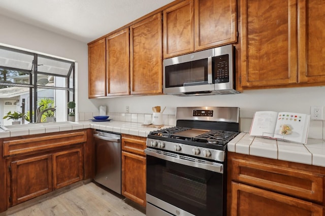 kitchen featuring a sink, brown cabinetry, tile countertops, light wood-style floors, and appliances with stainless steel finishes