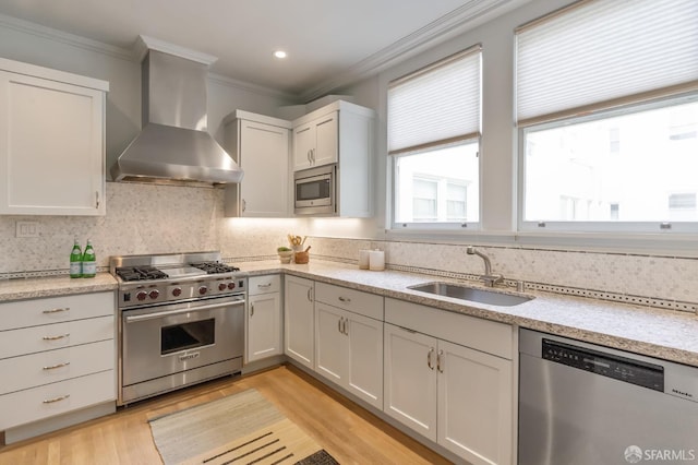 kitchen featuring light wood-type flooring, crown molding, wall chimney exhaust hood, appliances with stainless steel finishes, and a sink