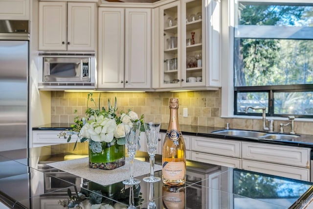 kitchen featuring glass insert cabinets, a sink, dark countertops, built in appliances, and backsplash