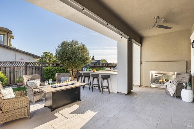 view of patio featuring an outdoor fire pit, a ceiling fan, fence, outdoor dry bar, and a glass covered fireplace