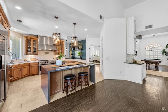 kitchen with glass insert cabinets, wall chimney range hood, stainless steel stove, a breakfast bar area, and backsplash