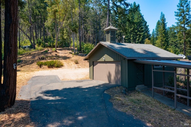 view of home's exterior with aphalt driveway, an outdoor structure, roof with shingles, a wooded view, and a garage