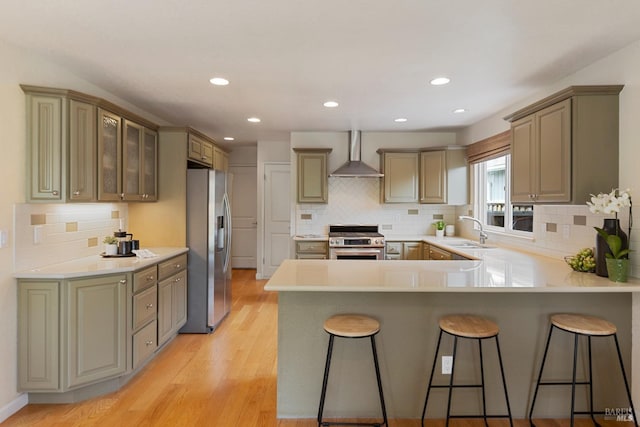 kitchen with wall chimney range hood, a peninsula, a kitchen bar, stainless steel appliances, and a sink