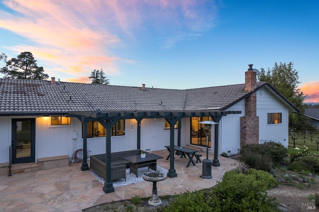 back of property at dusk with board and batten siding, outdoor lounge area, a patio, a tile roof, and a chimney
