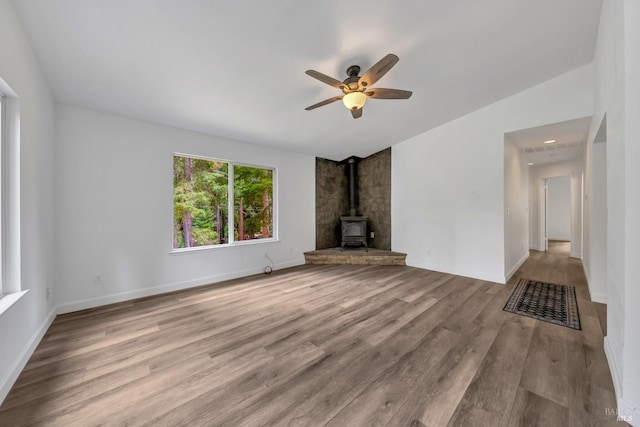 unfurnished living room featuring wood finished floors, a wood stove, a ceiling fan, and baseboards