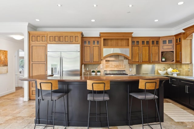 kitchen with an island with sink, under cabinet range hood, crown molding, tasteful backsplash, and stainless steel built in refrigerator