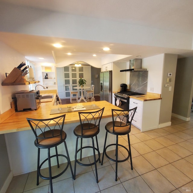 kitchen featuring wall chimney exhaust hood, a peninsula, stainless steel appliances, a sink, and butcher block counters