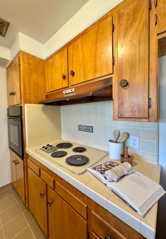 kitchen with white electric cooktop, under cabinet range hood, brown cabinetry, tasteful backsplash, and black oven