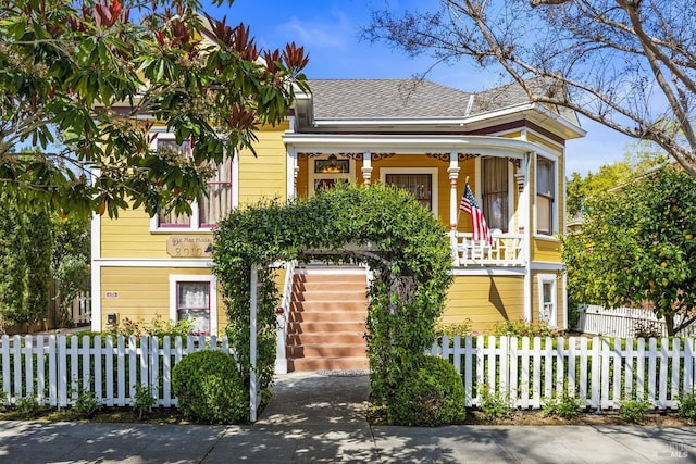 victorian-style house featuring roof with shingles, a porch, stairs, and a fenced front yard
