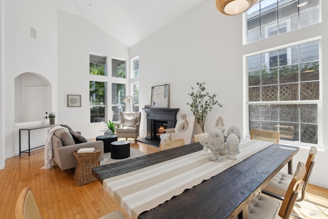 dining area with light wood finished floors, arched walkways, a glass covered fireplace, and high vaulted ceiling