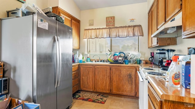 kitchen featuring freestanding refrigerator, under cabinet range hood, brown cabinets, electric stove, and a sink