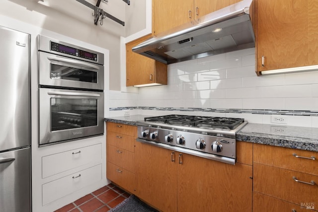 kitchen with under cabinet range hood, dark stone counters, decorative backsplash, stainless steel appliances, and dark tile patterned flooring