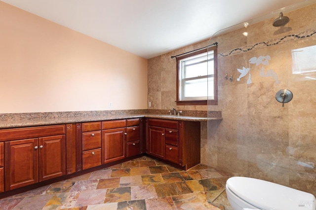 full bath featuring a tile shower, vanity, tile walls, stone finish flooring, and toilet