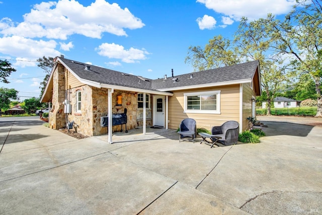 rear view of house with stone siding, a shingled roof, and a patio