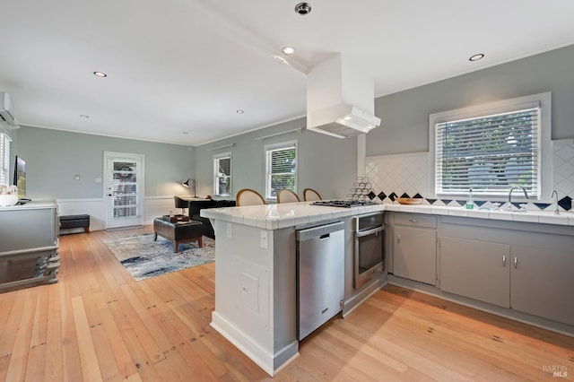 kitchen with wall chimney range hood, tile countertops, gray cabinets, a peninsula, and appliances with stainless steel finishes