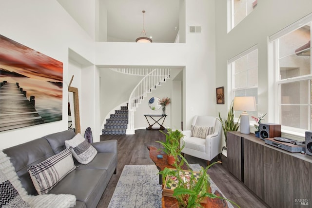living room with a healthy amount of sunlight, dark wood-style flooring, visible vents, a towering ceiling, and stairs