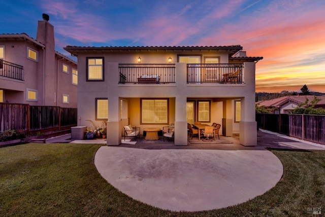 back of house at dusk featuring stucco siding, a balcony, a fenced backyard, and a lawn