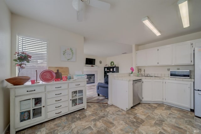 kitchen with stainless steel dishwasher, white cabinets, tile countertops, a glass covered fireplace, and stone finish flooring