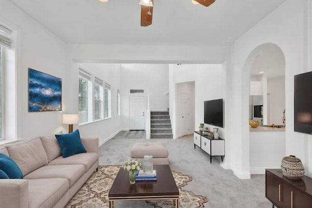 living room with stairway, a ceiling fan, baseboards, arched walkways, and light colored carpet