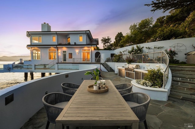 back of house at dusk featuring stairway, an outdoor kitchen, a chimney, a patio area, and outdoor dining area
