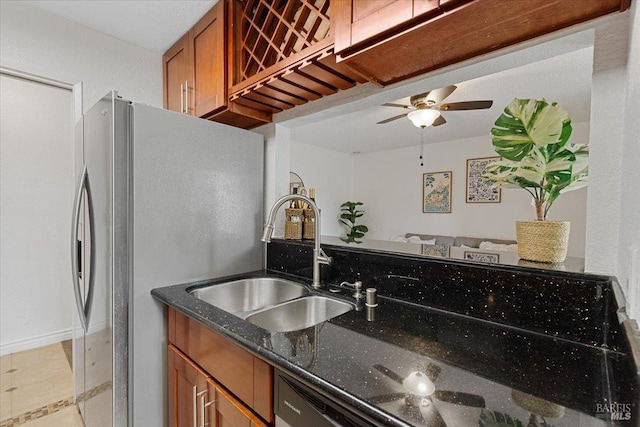 kitchen featuring ceiling fan, brown cabinetry, a sink, freestanding refrigerator, and dark stone counters