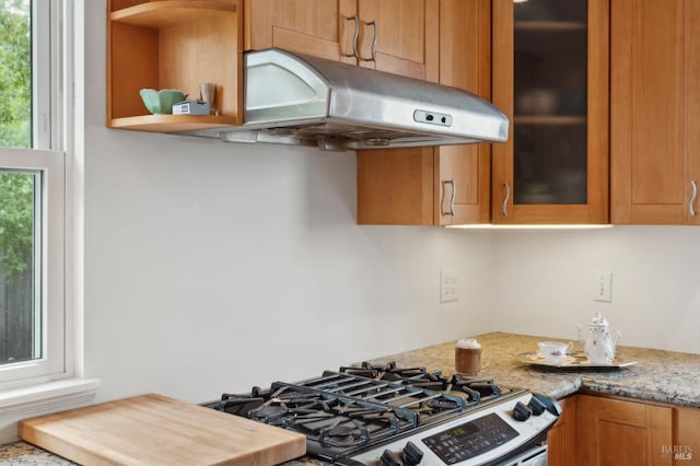 kitchen with stainless steel gas range, glass insert cabinets, light stone counters, brown cabinetry, and under cabinet range hood