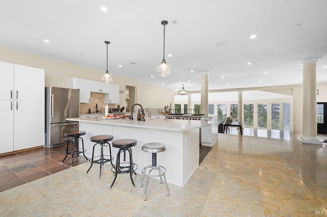 kitchen featuring freestanding refrigerator, ornate columns, an island with sink, white cabinets, and a breakfast bar