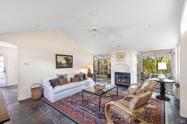 living room with stone tile floors, a ceiling fan, arched walkways, baseboards, and a glass covered fireplace