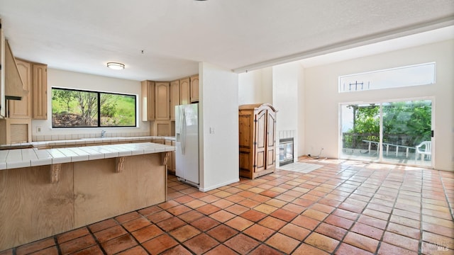 kitchen with light brown cabinets, tile counters, white refrigerator with ice dispenser, a breakfast bar area, and a peninsula
