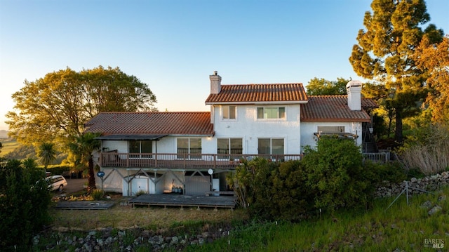 back of house at dusk with a wooden deck, a tile roof, a chimney, and stucco siding