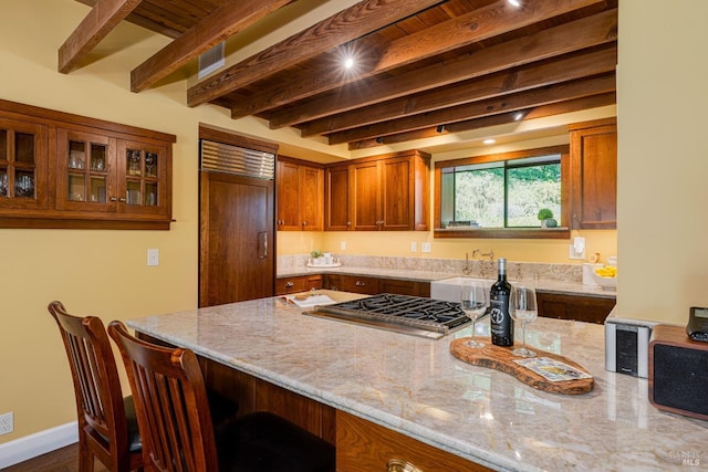 kitchen featuring stainless steel gas stovetop, a kitchen breakfast bar, light stone countertops, beam ceiling, and brown cabinetry