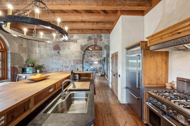 kitchen featuring premium appliances, wall chimney range hood, a sink, arched walkways, and beam ceiling